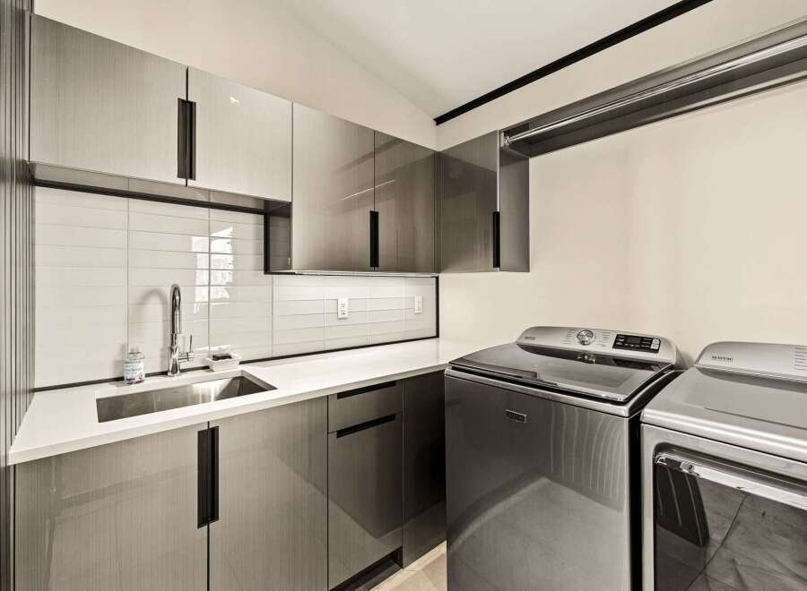 This modern laundry room features sleek, grey cabinetry with integrated handles, a white subway tile backsplash, and a stainless steel sink.