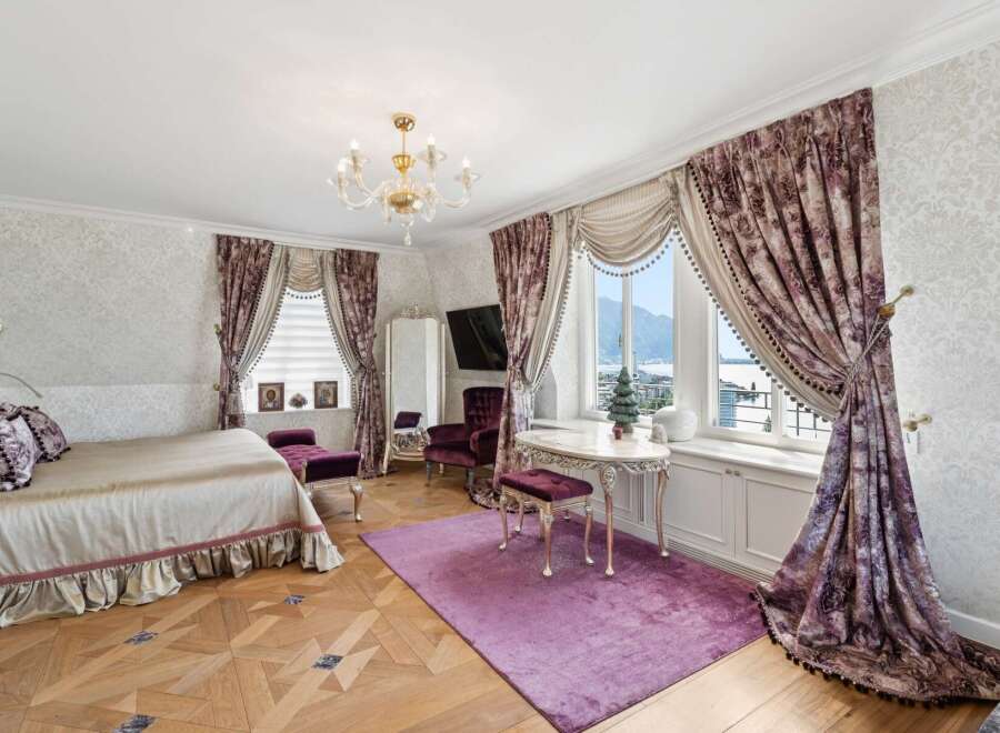 Experience opulent living in this luxurious bedroom featuring a grand bed with plush pillows and a ruffled skirt, complemented by rich, patterned curtains framing a stunning view.