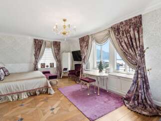 Experience opulent living in this luxurious bedroom featuring a grand bed with plush pillows and a ruffled skirt, complemented by rich, patterned curtains framing a stunning view.