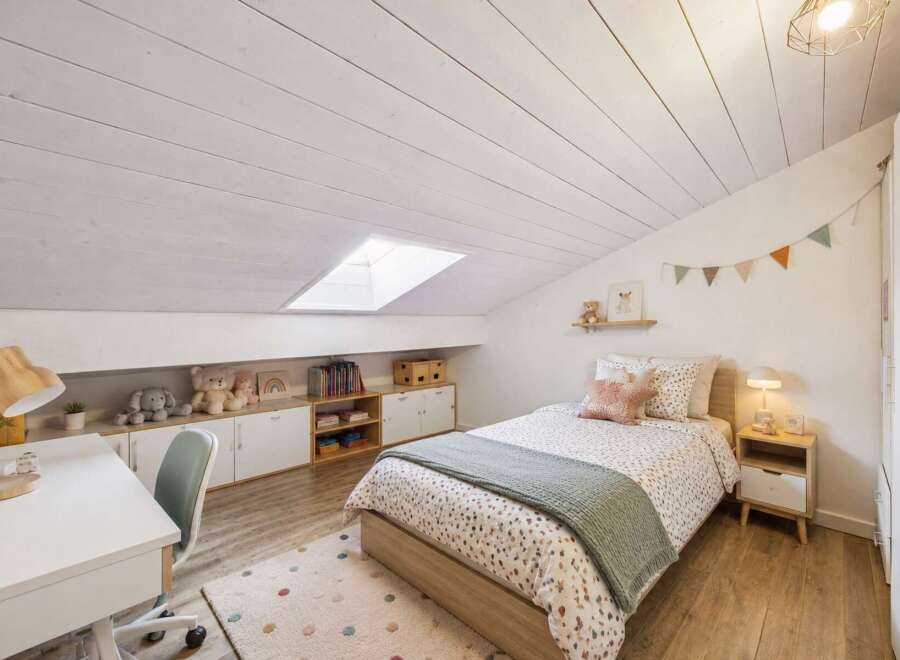 This charming attic bedroom features a cozy bed with playful bedding, a functional desk area with a wooden lamp, and ample storage solutions.