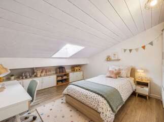 This charming attic bedroom features a cozy bed with playful bedding, a functional desk area with a wooden lamp, and ample storage solutions.