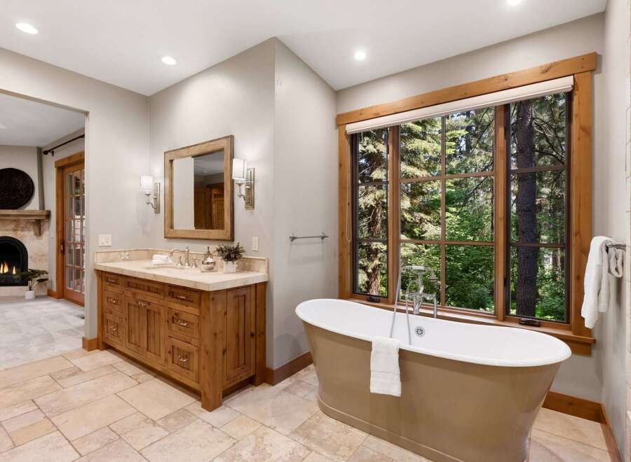 Indulge in luxury with this stunning bathroom featuring a freestanding soaking tub perfectly positioned to enjoy breathtaking forest views through a large, wood-trimmed window.