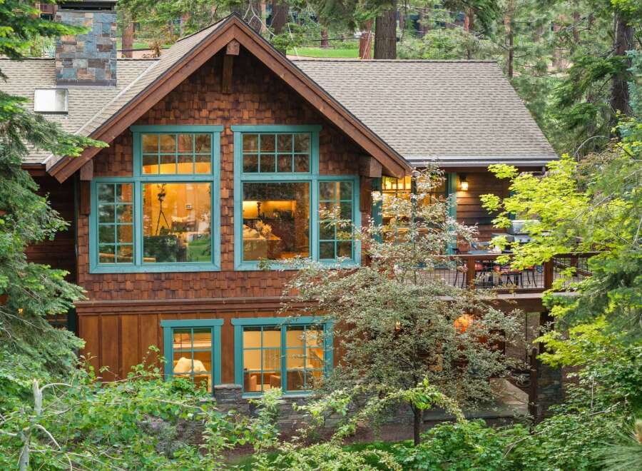 Nestled amidst lush greenery, this charming wooden home features rustic cedar shake siding and striking teal-trimmed windows that add a pop of color.