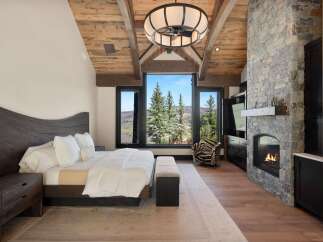 Experience luxury mountain living in this stunning bedroom featuring a grand stone fireplace and expansive windows with breathtaking views.