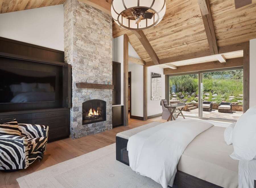 Experience luxury mountain living in this stunning bedroom featuring a grand stone fireplace and a vaulted wood-beam ceiling.