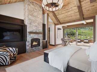 Experience luxury mountain living in this stunning bedroom featuring a grand stone fireplace and a vaulted wood-beam ceiling.