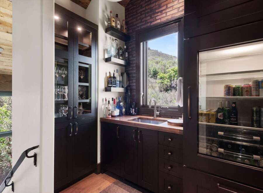 This stunning home bar features a dark wood cabinet with glass doors displaying elegant glassware, complemented by a brick accent wall and a large window offering scenic views.
