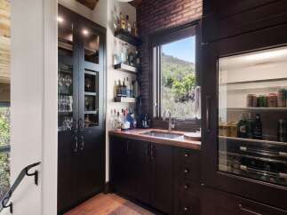 This stunning home bar features a dark wood cabinet with glass doors displaying elegant glassware, complemented by a brick accent wall and a large window offering scenic views.