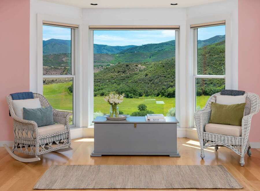 Experience breathtaking mountain views from this charming room, featuring a cozy seating area with two wicker chairs and a large bay window.