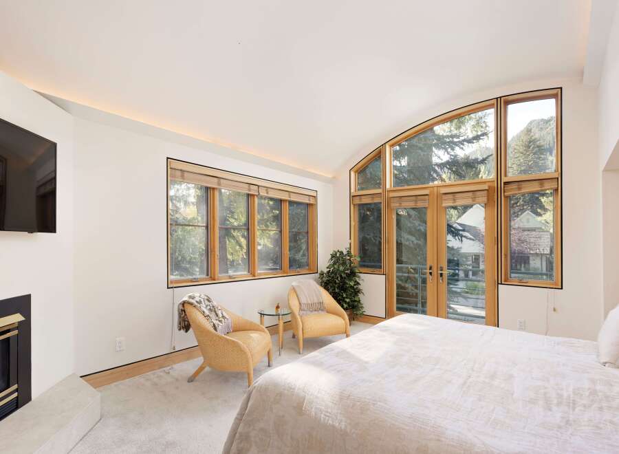 Experience luxury mountain living in this stunning bedroom featuring a cozy fireplace and a large, arched window offering breathtaking views of the surrounding trees and mountains.