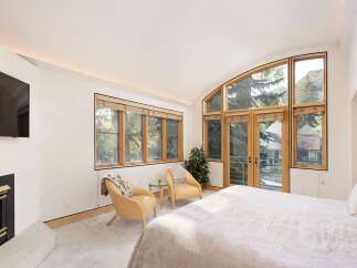 Experience luxury mountain living in this stunning bedroom featuring a cozy fireplace and a large, arched window offering breathtaking views of the surrounding trees and mountains.