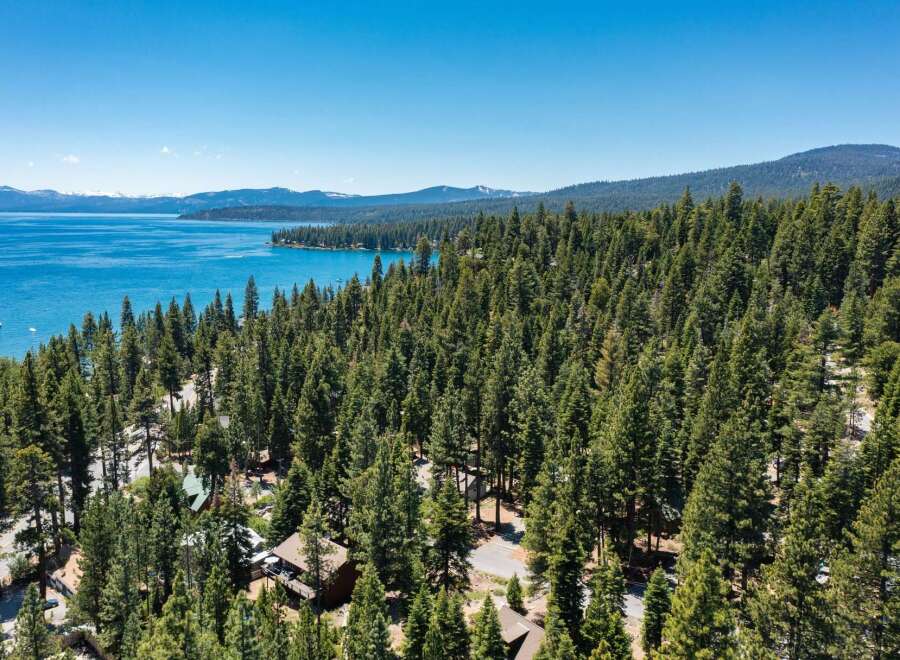 Experience the ultimate lakeside living with this stunning aerial view of a property nestled amidst a dense pine forest, offering breathtaking vistas of a crystal-clear blue lake.