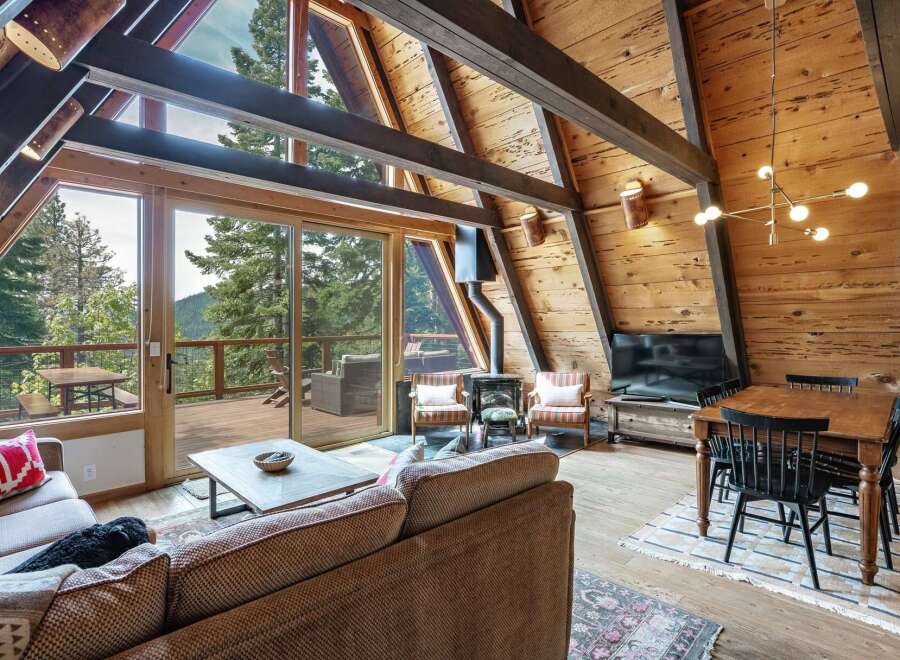Experience rustic charm in this A-frame cabin living room, featuring a stunning wood-paneled interior with exposed beams and a wall of windows offering breathtaking mountain views.