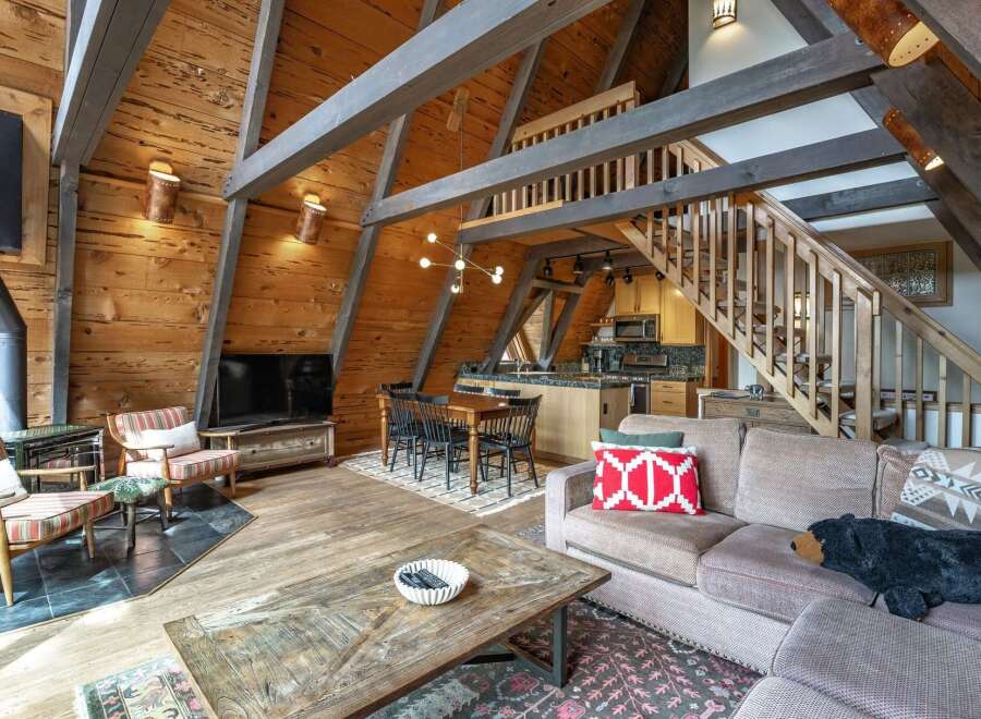 Experience rustic charm in this A-frame cabin living space, featuring a cozy fireplace, a well-equipped kitchen, and a dining area perfect for gathering.