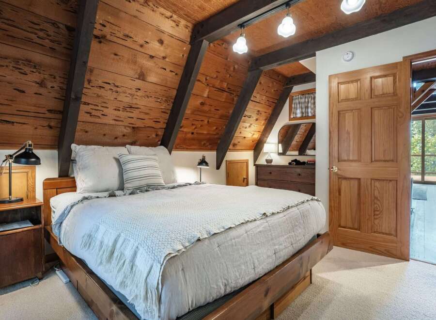 Experience rustic charm in this cozy bedroom featuring a wooden plank A-frame ceiling with exposed beams and a comfortable bed adorned with plush linens.