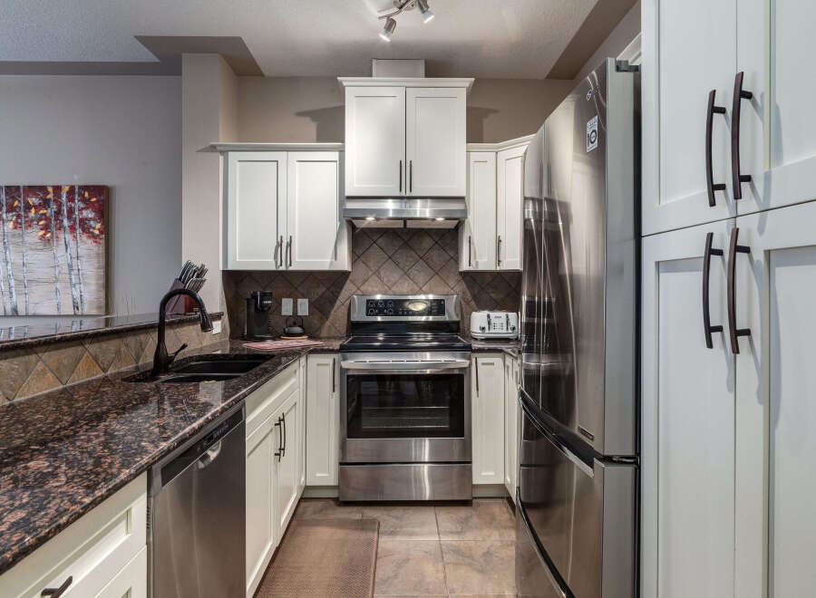 This modern kitchen boasts sleek stainless steel appliances, including a refrigerator, oven, and dishwasher, complemented by elegant white cabinetry and granite countertops.