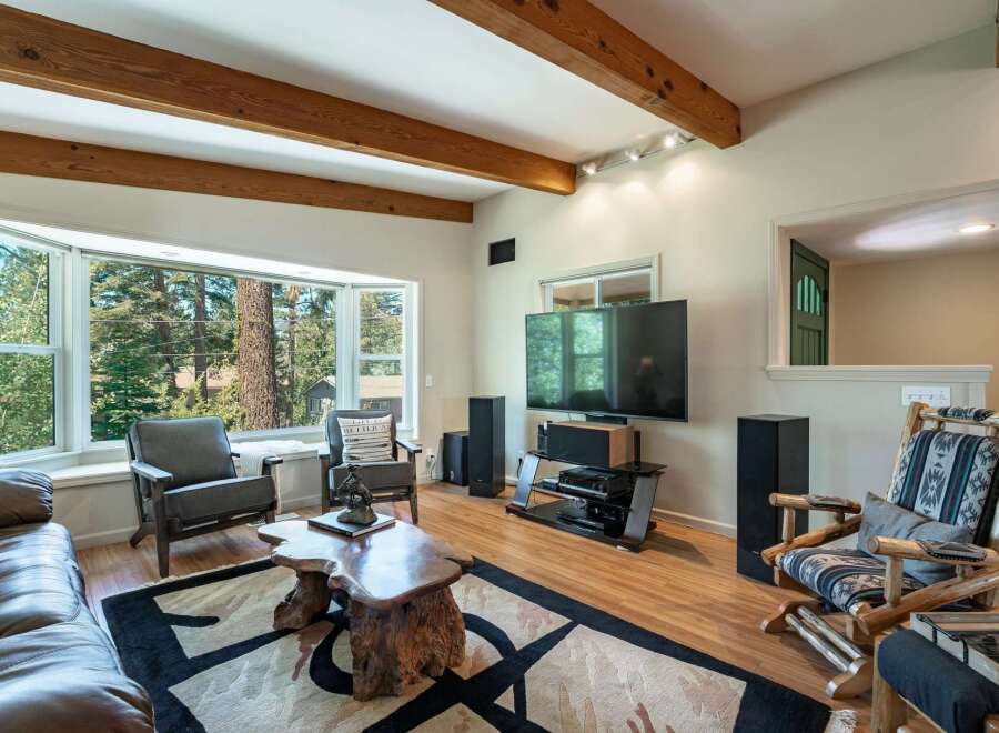 Experience cozy living in this inviting living room, featuring a large bay window with stunning forest views and exposed wooden beams on the ceiling.