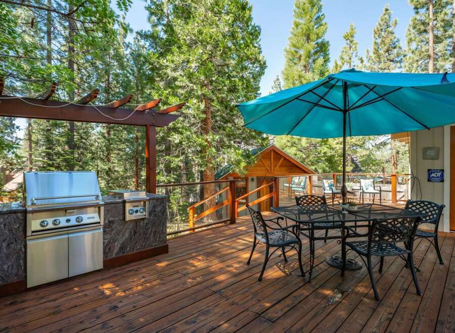 Enjoy outdoor living at its finest on this spacious wooden deck, complete with a built-in stainless steel grill and a large turquoise umbrella providing shade over a dining set.