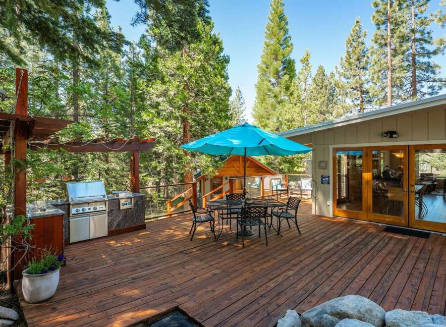Enjoy outdoor living at its finest on this spacious wooden deck, complete with a built-in grill station and a charming dining set under a vibrant blue umbrella.