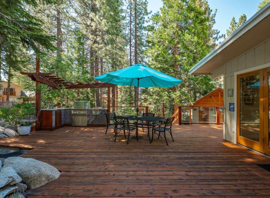 Enjoy outdoor living at its finest on this expansive wooden deck, complete with a built-in outdoor kitchen and a charming dining set under a vibrant turquoise umbrella.