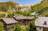 Experience alpine living in this stunning mountain chalet, featuring rustic stone and timber construction set against a breathtaking backdrop of snow-capped peaks and lush green slopes.