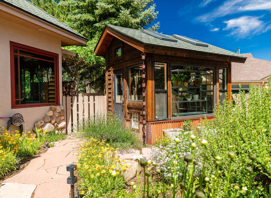 Discover this charming property featuring a rustic wooden greenhouse, perfect for gardening enthusiasts, alongside a cozy home with a distinctive red-trimmed window.