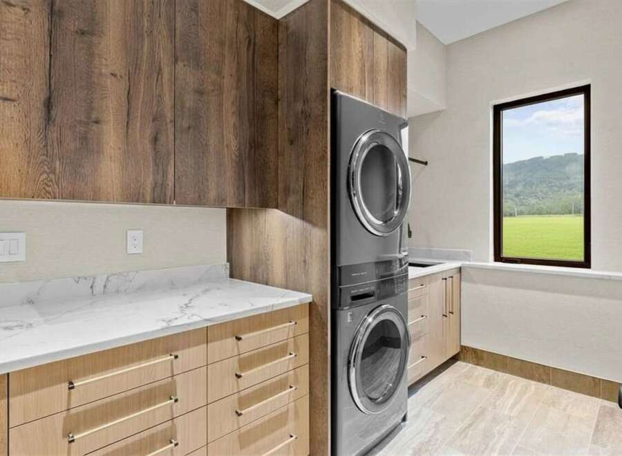 This modern laundry room boasts a stacked washer and dryer unit, complemented by sleek wood cabinetry and elegant marble countertops.
