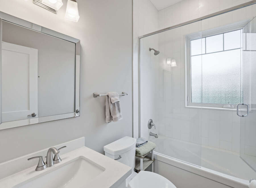 This bright and modern bathroom features a sleek vanity with a silver faucet, a clean white toilet, and a spacious bathtub with a glass enclosure.
