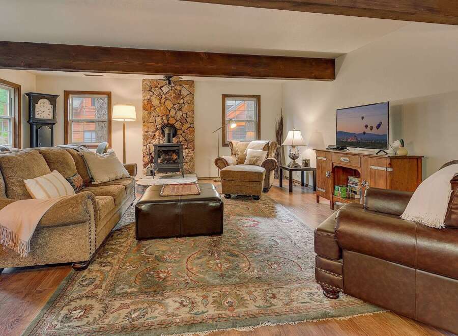 Cozy living room featuring a stone fireplace with a wood-burning stove, comfortable seating including a plush sofa and armchair, and a large area rug.