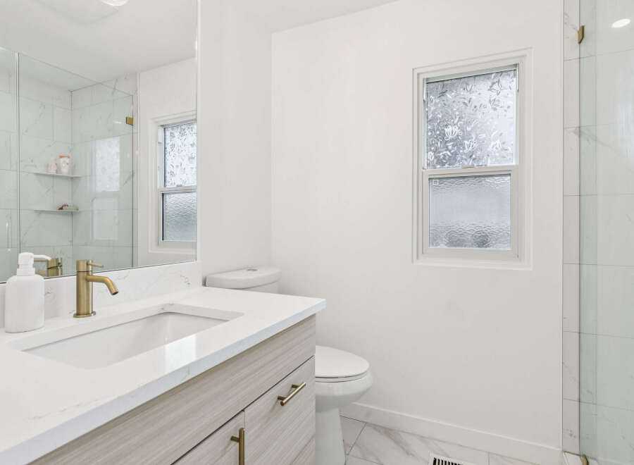 This modern bathroom features a sleek vanity with a quartz countertop and gold fixtures, complemented by a large mirror and a spacious glass-enclosed shower with marble tile.