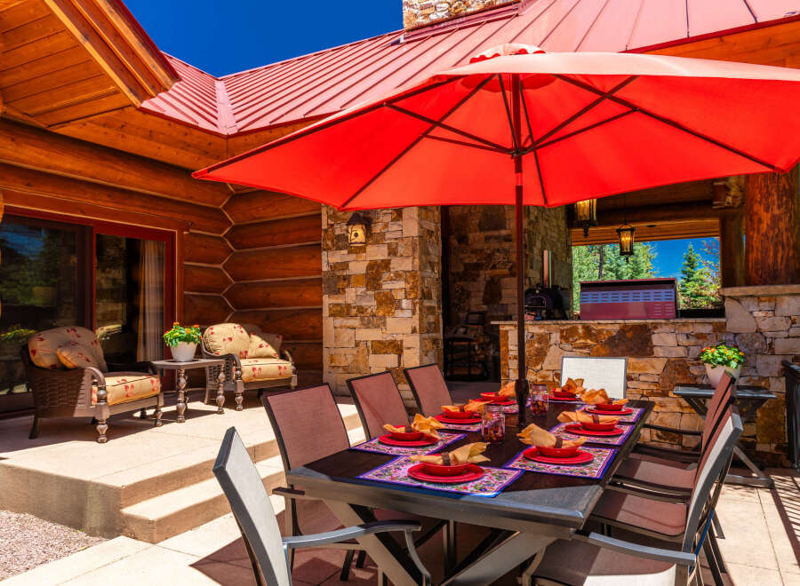 Enjoy al fresco dining on this stunning mountain property with a spacious outdoor patio, complete with a large dining table set for guests and a vibrant red umbrella for shade.