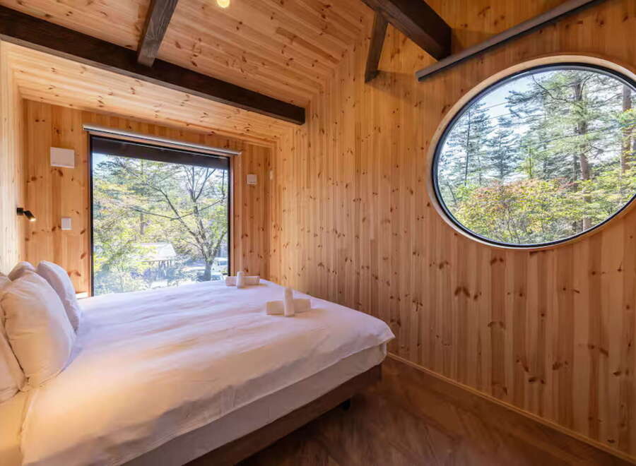 Experience serene living in this charming wooden cabin, featuring a comfortable bed bathed in natural light from a large picture window overlooking a lush forest.