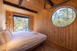 Experience serene living in this charming wooden cabin, featuring a comfortable bed bathed in natural light from a large picture window overlooking a lush forest.