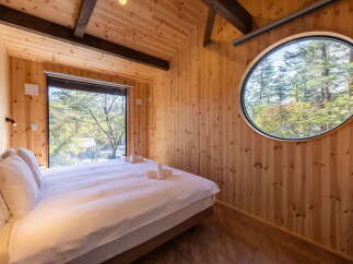 Experience serene living in this charming wooden cabin, featuring a comfortable bed bathed in natural light from a large picture window overlooking a lush forest.