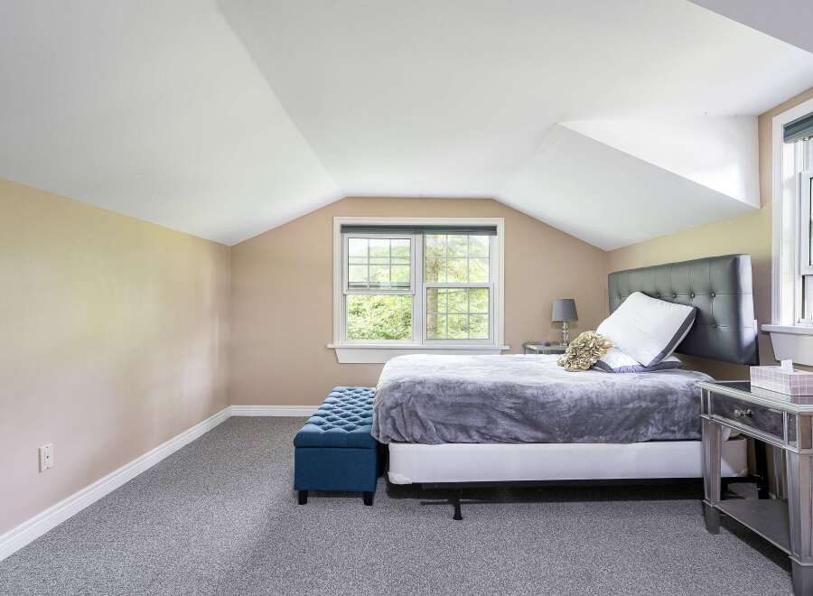 Discover comfort and style in this charming bedroom, featuring a plush grey comforter on a queen-sized bed and a sophisticated tufted blue ottoman.