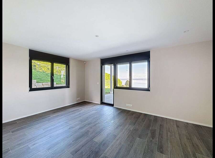 Experience modern living in this spacious room featuring large windows that offer stunning views of a lush green hillside and a serene lake.