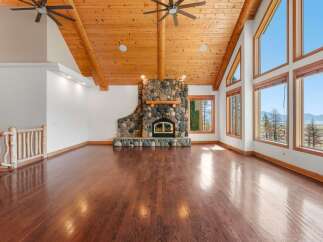 Experience breathtaking lake and mountain views from this stunning living room, featuring a grand stone fireplace and soaring vaulted ceilings with exposed wooden beams.