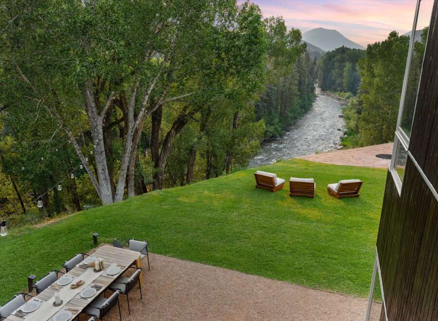 Experience luxurious outdoor living with this stunning property featuring a spacious dining area and a serene seating space overlooking a picturesque river and mountain landscape.