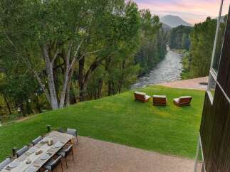 Experience luxurious outdoor living with this stunning property featuring a spacious dining area and a serene seating space overlooking a picturesque river and mountain landscape.