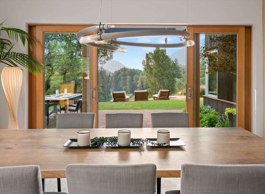 Experience seamless indoor-outdoor living with this stunning dining area featuring a large wooden table and chairs, opening up to a breathtaking mountain view.