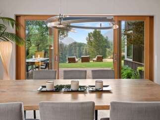 Experience seamless indoor-outdoor living with this stunning dining area featuring a large wooden table and chairs, opening up to a breathtaking mountain view.