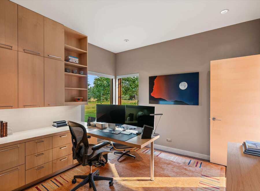 This modern home office features a sleek built-in wooden cabinetry system with ample storage, complemented by a spacious desk equipped with dual monitors.