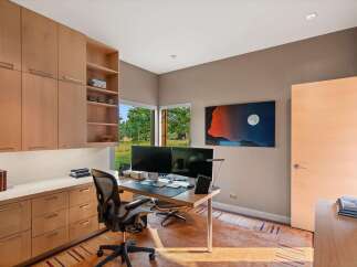 This modern home office features a sleek built-in wooden cabinetry system with ample storage, complemented by a spacious desk equipped with dual monitors.