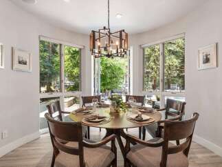 Dine in style in this elegant dining room, featuring a round wooden table set for six and surrounded by large windows that offer a lush green view.