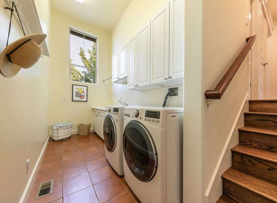 This spacious laundry room features a side-by-side washer and dryer set, ample white cabinetry for storage, and a large window offering natural light.