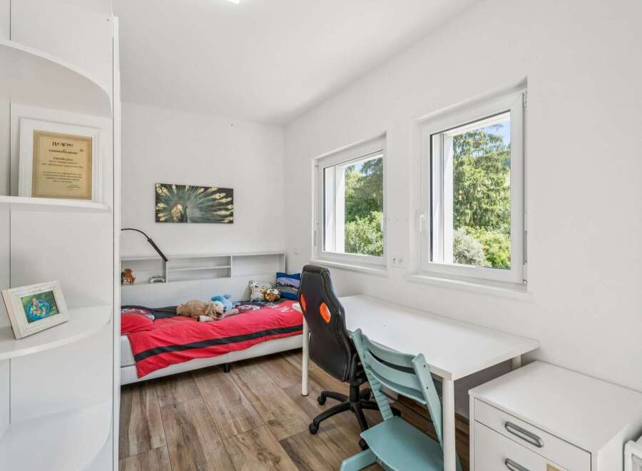 This bright and airy bedroom features a comfortable bed with red bedding, a dedicated study area with a white desk and ergonomic chairs, and ample natural light from two large windows.