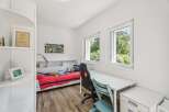 This bright and airy bedroom features a comfortable bed with red bedding, a dedicated study area with a white desk and ergonomic chairs, and ample natural light from two large windows.