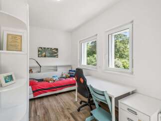 This bright and airy bedroom features a comfortable bed with red bedding, a dedicated study area with a white desk and ergonomic chairs, and ample natural light from two large windows.