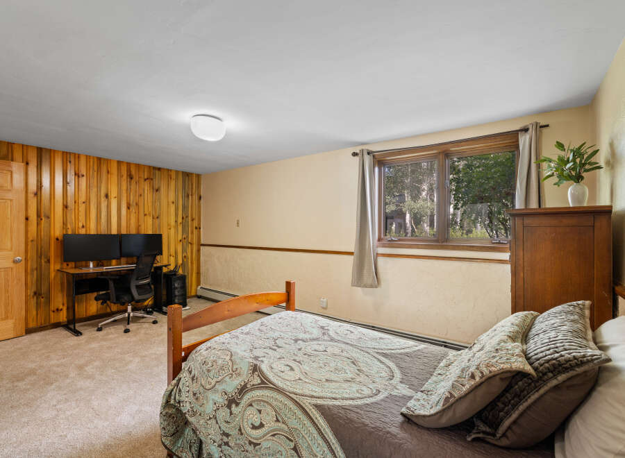 This cozy bedroom features a comfortable bed with decorative pillows, a functional workspace with dual monitors, and a charming wooden dresser.