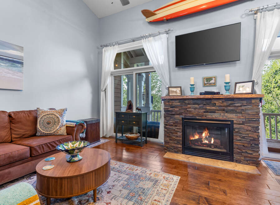 Cozy living room featuring a warm brown leather sofa, a rustic stone fireplace with a roaring fire, and a large flat-screen TV mounted above.